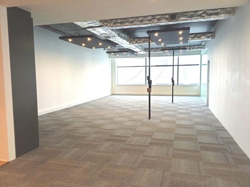 A spacious, empty open-plan office area featuring exposed industrial ceilings, carpeted flooring, and large windows providing ample natural light.