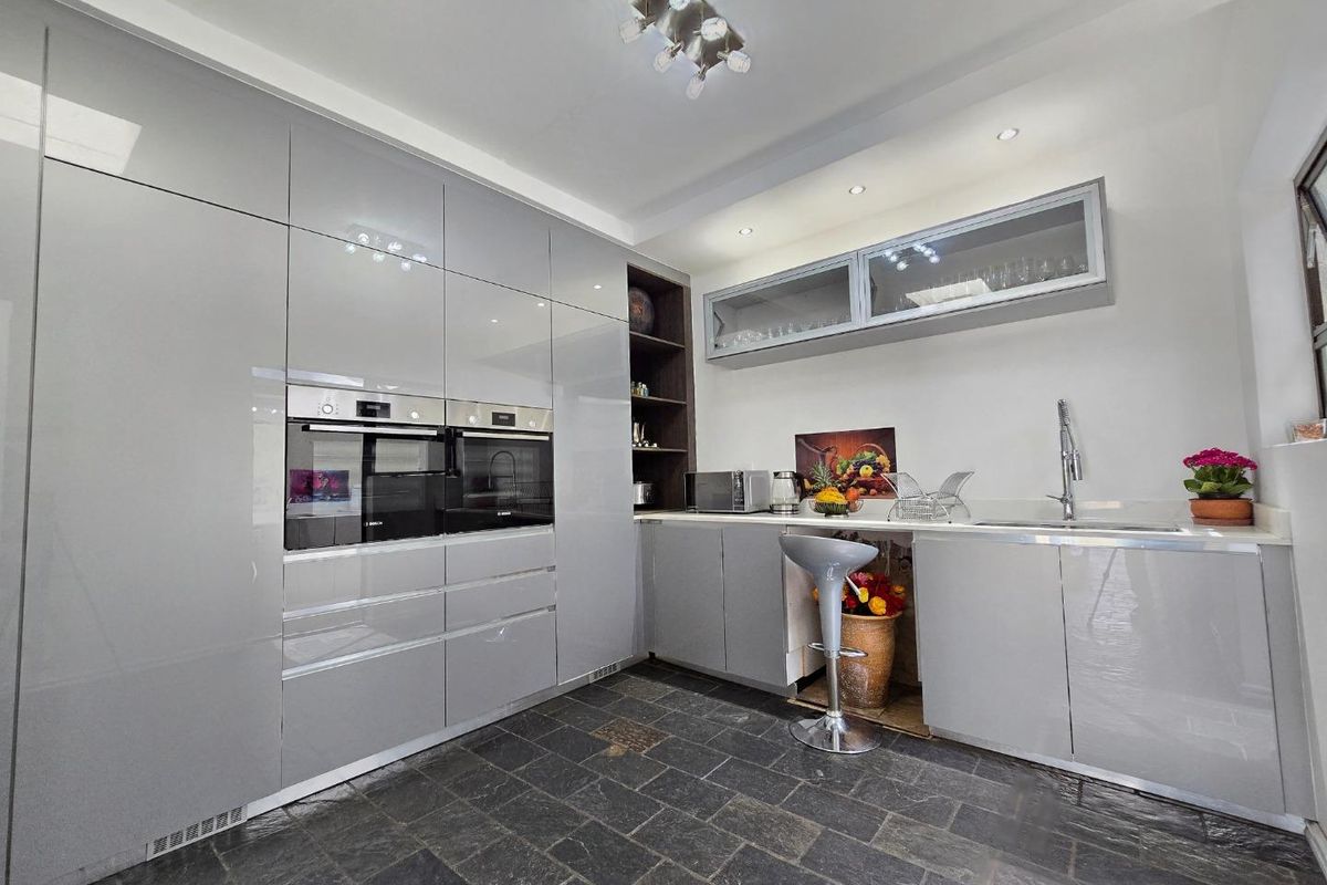 Modern kitchen featuring high-gloss grey cabinetry, built-in double ovens, stone-style floor tiling, and integrated sink with overhead glass-fronted storage cabinets.