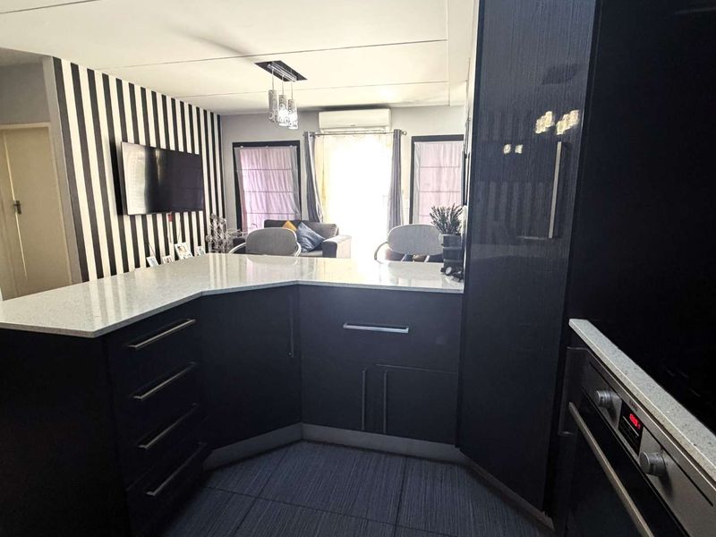 Modern open-plan kitchen featuring dark cabinetry, stone countertops, and integrated oven, flowing into a living area with tiled floors and air conditioning.