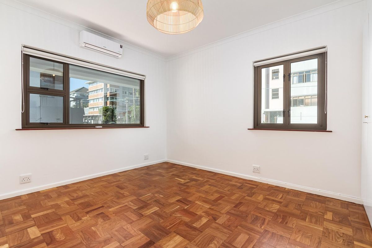 A bright, empty bedroom featuring polished parquet flooring, a wall-mounted split-system air conditioner, and large windows with dark aluminium frames.