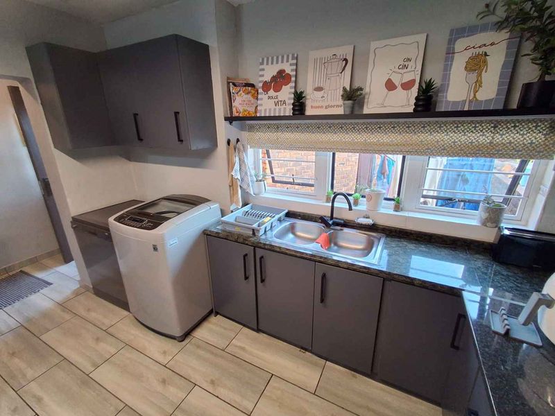 A functional laundry area featuring modern grey cabinetry, granite countertops, a double basin sink, and tiled flooring with window security bars.