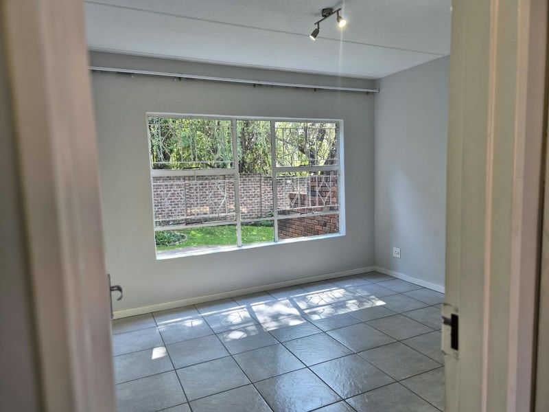 A bright, empty bedroom featuring neutral tiled flooring, track lighting, and a large window with security bars overlooking a garden area.