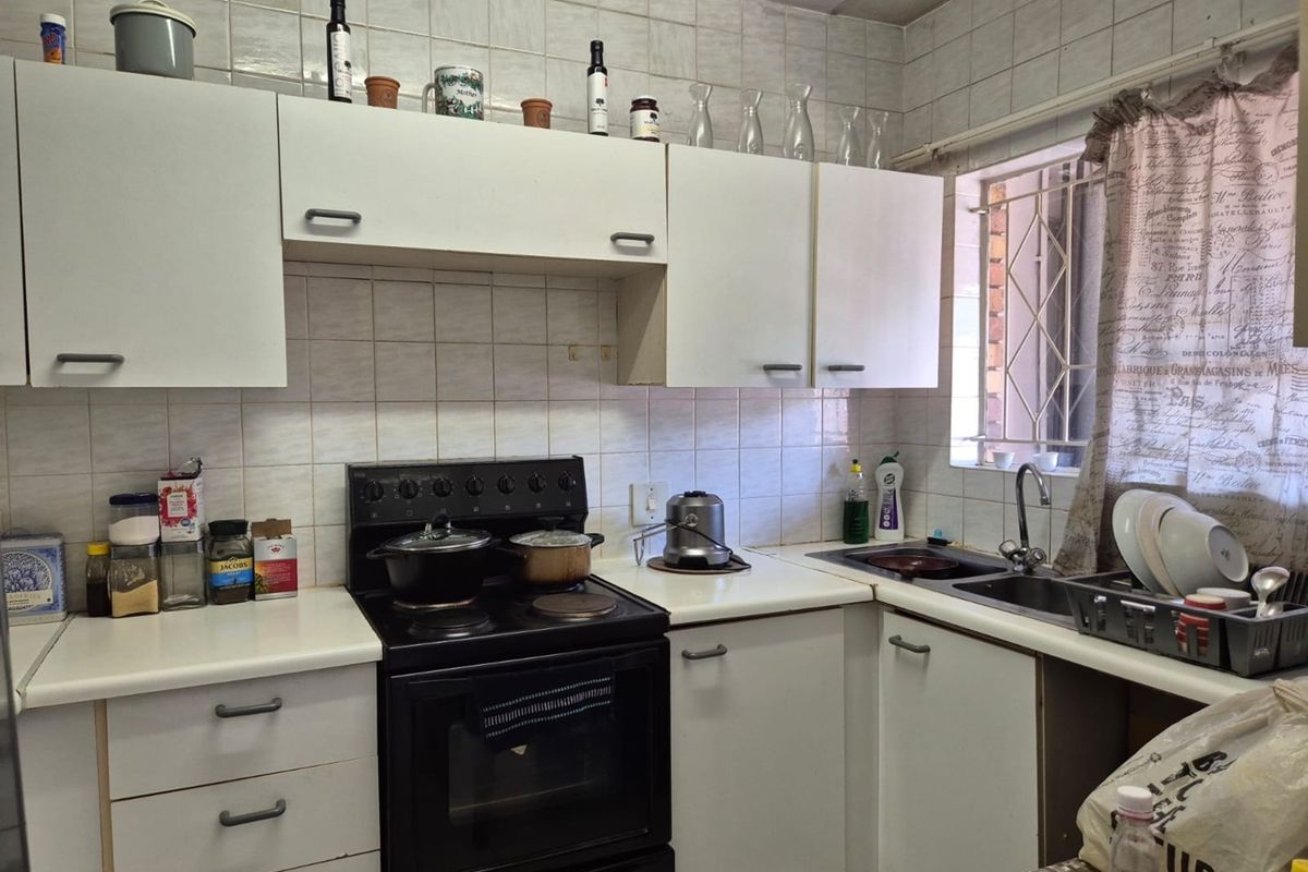 A functional kitchen featuring white cabinetry, laminate countertops, a stainless steel sink, and a freestanding stove, with tiled walls and a window security gate.