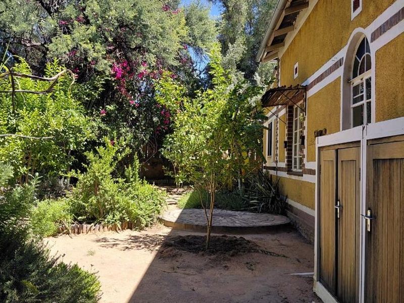 A rear garden area featuring a textured exterior wall, arched window, timber doors, and a small stone paved section surrounded by mature trees.