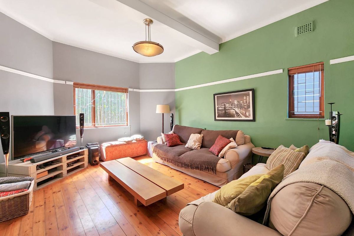 A cozy living room featuring original hardwood flooring, picture rails, and large windows fitted with security bars.