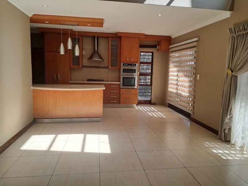 Spacious open-plan kitchen and living area featuring warm-toned wooden cabinetry, built-in appliances, tiled flooring, and large windows with modern roller blinds.
