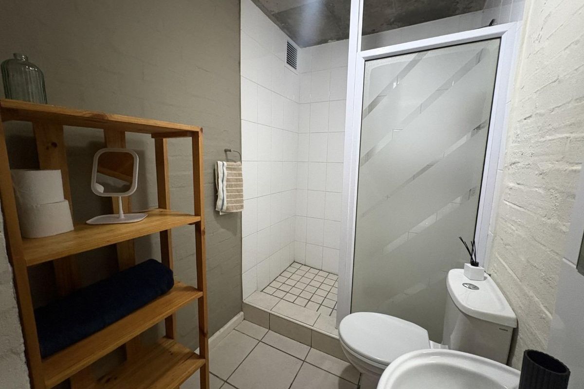 A compact bathroom featuring a walk-in shower with white tiling, a standard toilet, and a pedestal basin with industrial-style exposed concrete ceiling.