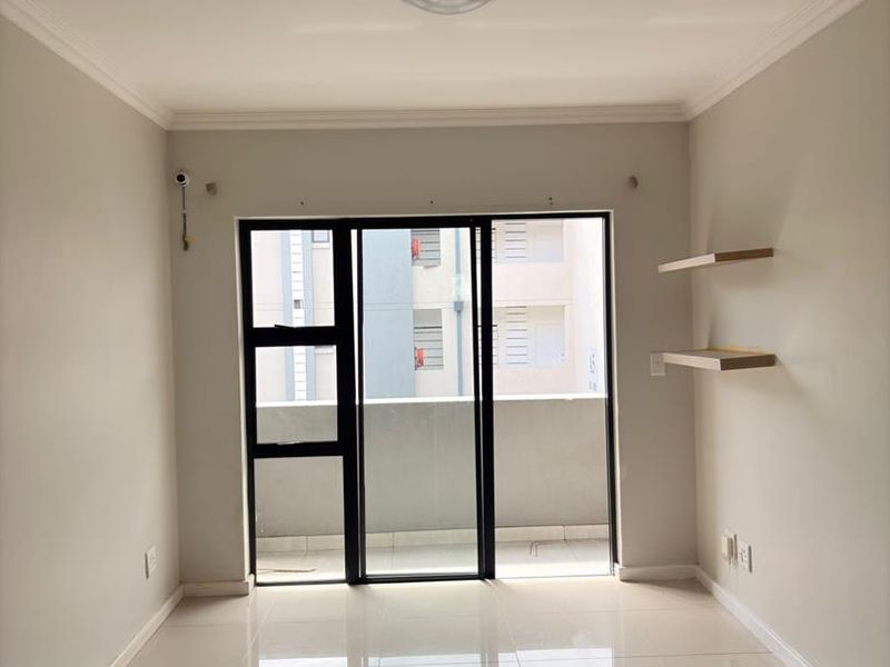 A bright, empty living room featuring neutral tiled flooring, modern sliding glass doors leading to a balcony, and minimalist floating wall shelves.