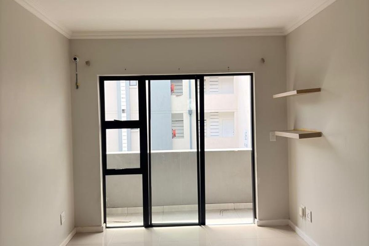 A bright, empty living room featuring neutral tiled flooring, modern sliding glass doors leading to a balcony, and minimalist floating wall shelves.