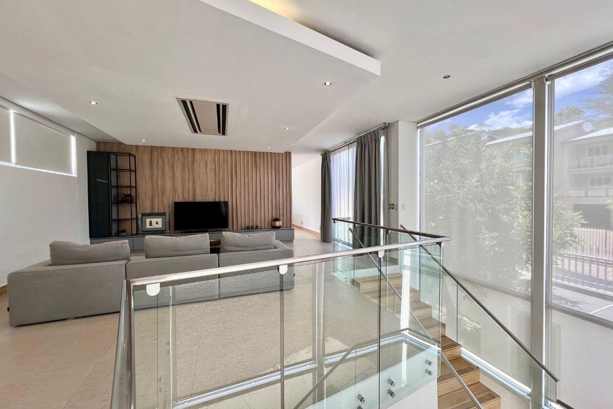 Modern open-plan living area featuring a wood-paneled feature wall, recessed ceiling lighting, ducted air conditioning, and a glass-balustrade staircase.