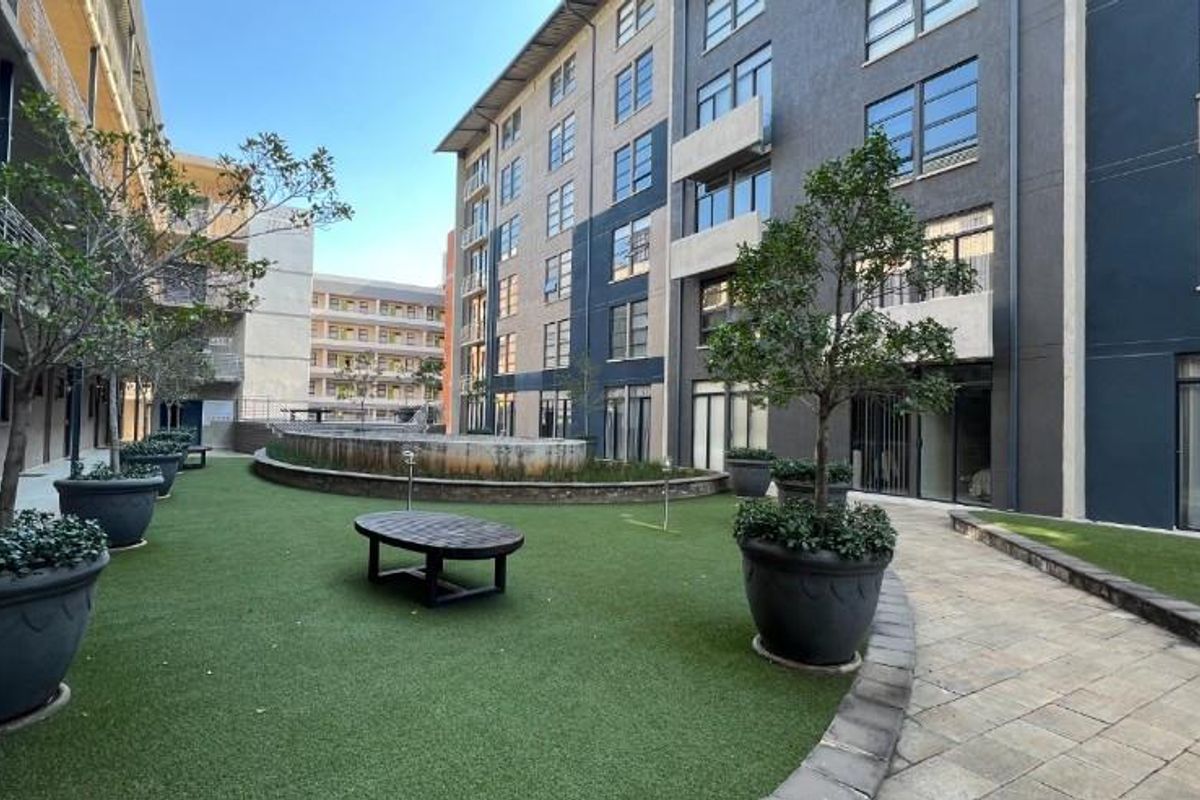 A well-maintained communal courtyard area featuring artificial lawn, paved pathways, and a raised garden bed set against a modern multi-story apartment building.