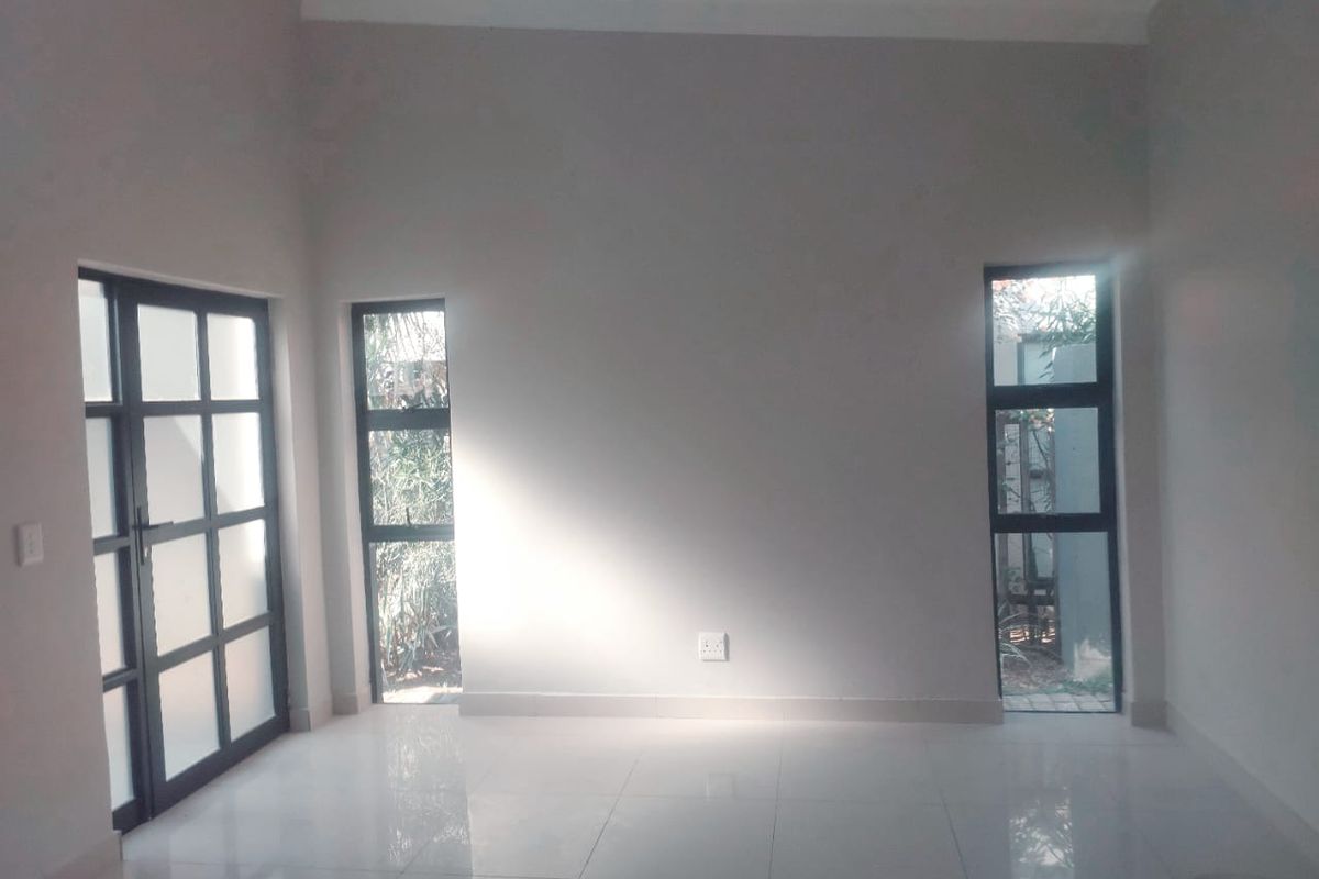 A bright, empty living room featuring neutral tiled flooring, modern aluminium-framed windows, and a matching glass door leading to the exterior.