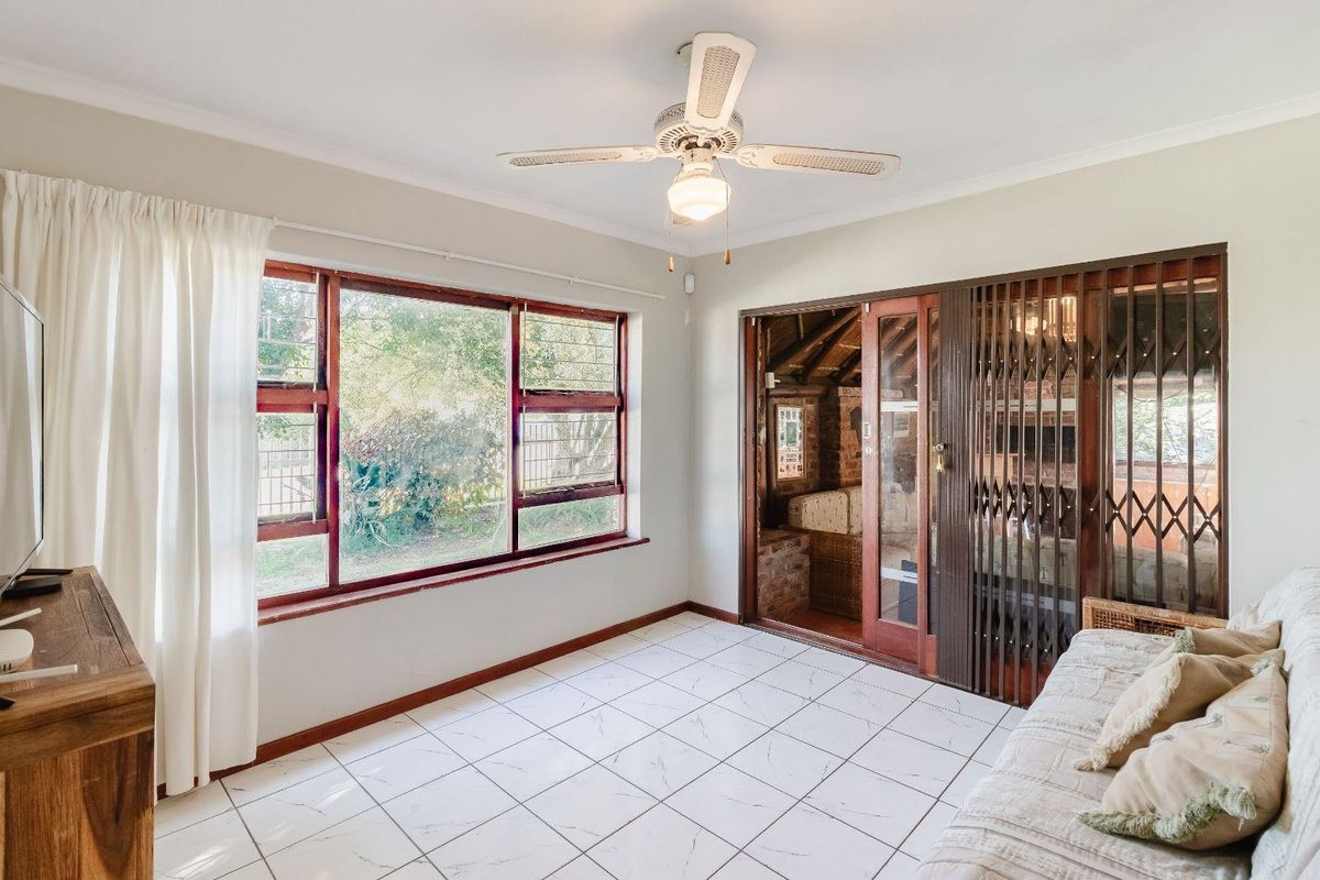 A sunlit living room featuring tiled flooring, a ceiling fan, and large windows, with sliding door access to an adjacent covered patio area.