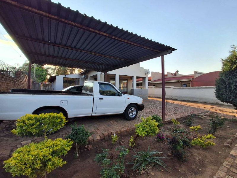 Front exterior view featuring a paved driveway, a corrugated metal carport, and established garden beds leading to the main house entrance.