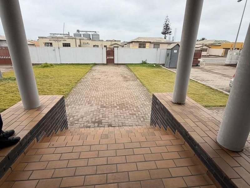 View from a covered porch overlooking a paved driveway, lawn, and perimeter wall in an office property setting.