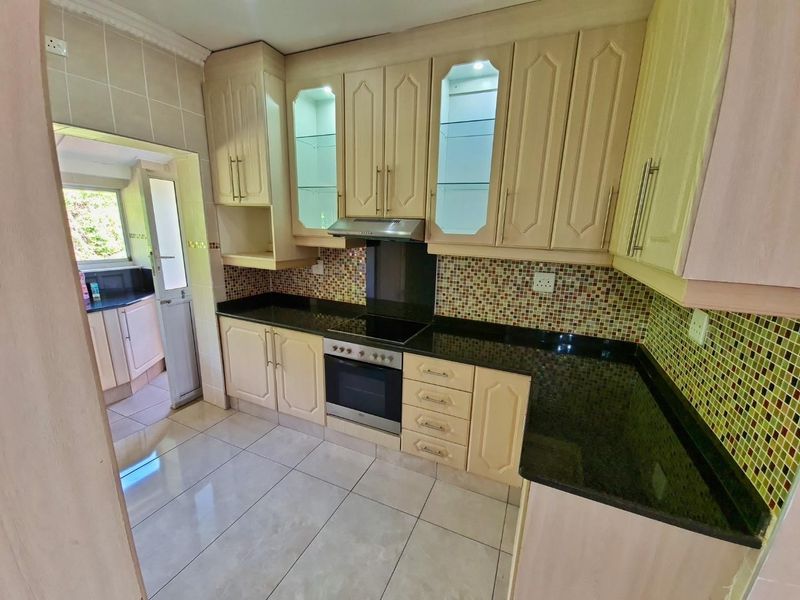 A well-maintained kitchen featuring light-toned cabinetry, granite countertops, a mosaic tile backsplash, and integrated appliances, with an adjacent scullery area.