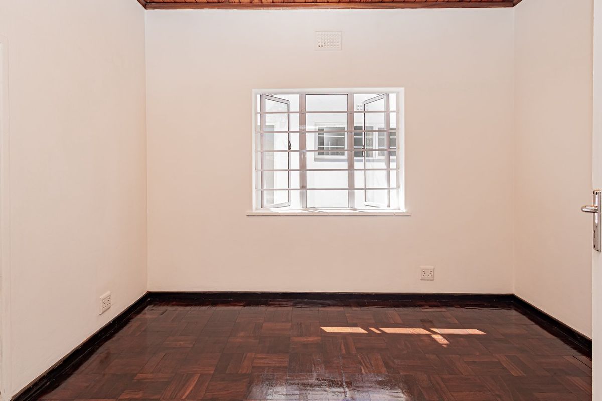 A bright, empty bedroom featuring polished parquet flooring, a timber ceiling trim, and a window fitted with security bars.