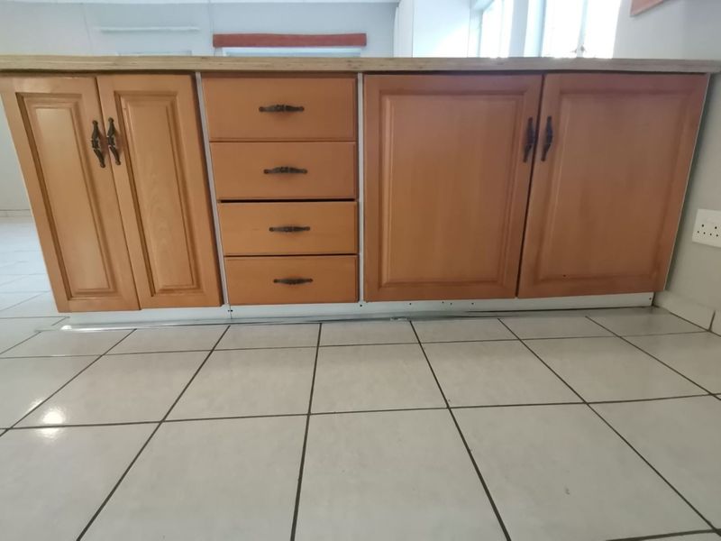 A close-up view of dated wooden kitchen cabinetry with a laminate countertop and ceramic floor tiles, showing signs of wear and missing kickplate trim.