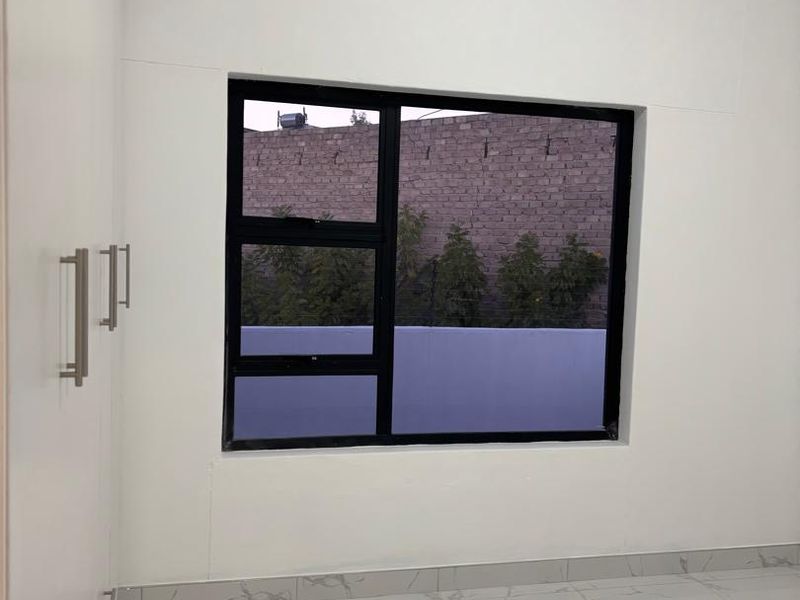 A bright, empty bedroom featuring modern white tiled flooring, built-in cupboards, and a large aluminium-framed window.