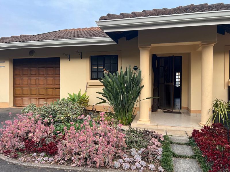 Front elevation of a townhouse featuring a covered entrance with columns, a tiled roof, and a single garage door, complemented by a landscaped garden.