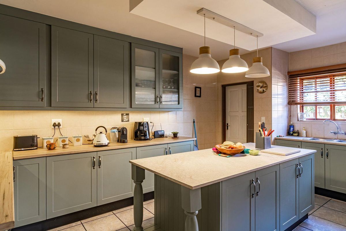 A well-maintained kitchen featuring sage green cabinetry, a central island with stone countertop, tiled backsplash, and modern pendant lighting.