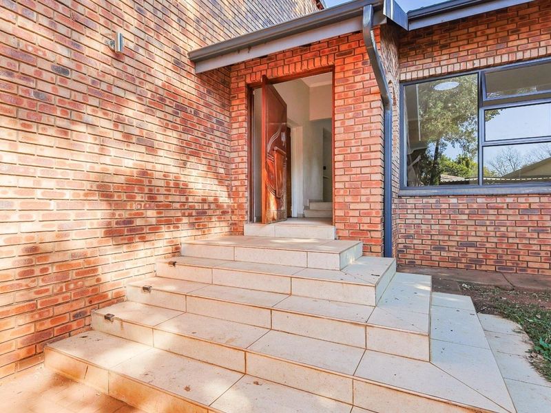 A well-maintained front entrance featuring a classic face brick facade, tiled steps leading to a solid timber front door, and modern aluminium window frames.