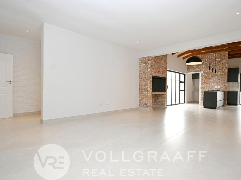 Spacious open-plan living area featuring exposed brick accents, timber ceiling beams, tiled flooring, and a modern kitchen with built-in cabinetry.