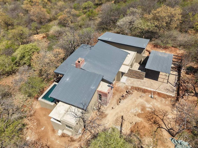 Aerial view of a modern house featuring a metal roof, brick chimney, private swimming pool, and a carport, set within a natural bushveld environment.