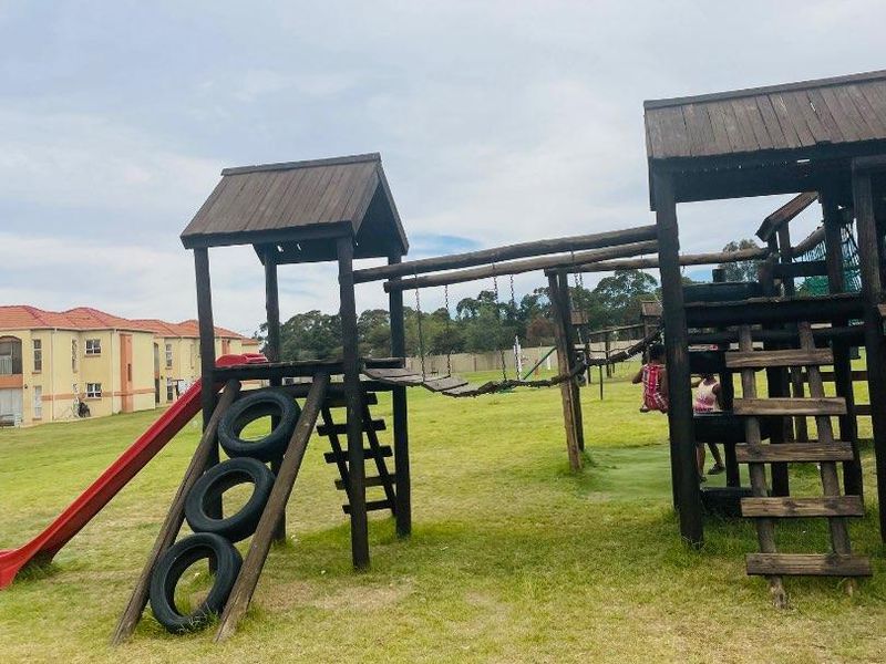 Outdoor communal playground area featuring wooden climbing structures, a slide, and a bridge, situated on a grassed common area within the apartment complex.