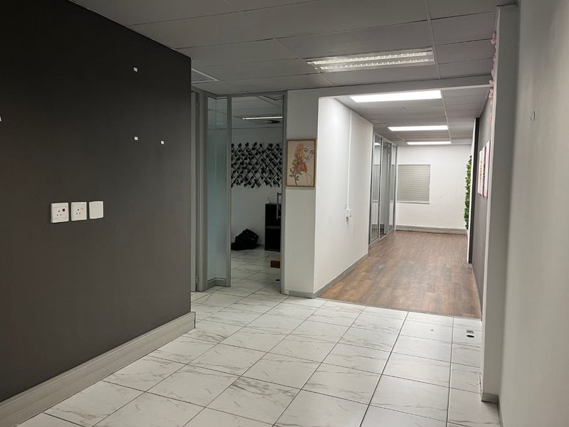 A spacious office hallway featuring a mix of tiled and wood-look flooring, glass partitions, and recessed ceiling lighting, offering a clean, professional layout.