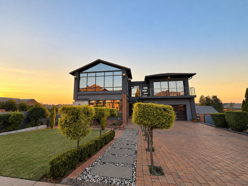 Modern double-story residence featuring a paved driveway, landscaped garden, and expansive glass windows, showcasing a contemporary architectural design at sunset.