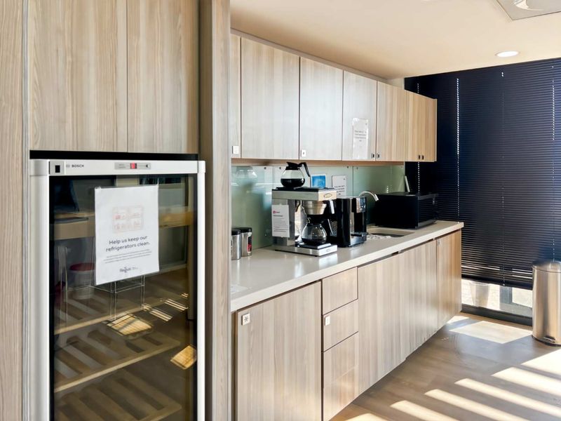 A modern office kitchenette featuring light wood-grain cabinetry, integrated refrigerator, sleek countertops, and a sink, complemented by natural light through venetian blinds.