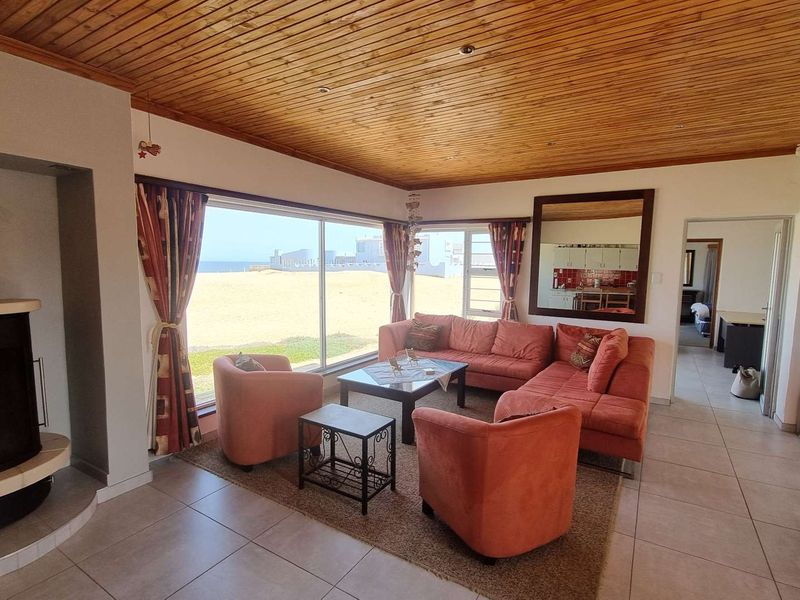 Spacious living room featuring a wood-paneled ceiling, tiled floors, and a freestanding fireplace, with large windows offering direct views of the beach.
