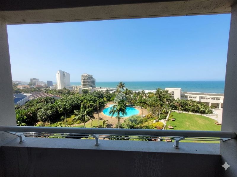 A balcony view overlooking a landscaped garden, swimming pool, and the ocean in the distance.