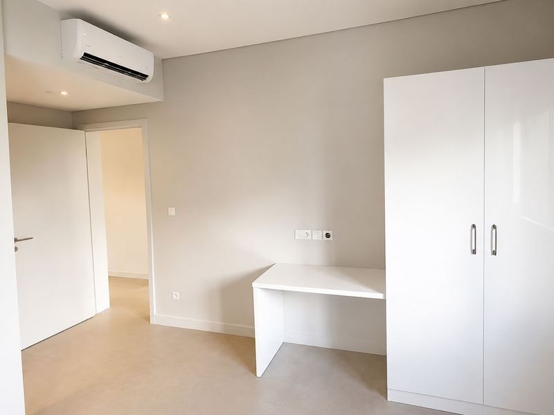 A modern, neutral-toned bedroom featuring a built-in wardrobe, integrated desk, split-system air conditioning, and clean tiled flooring.