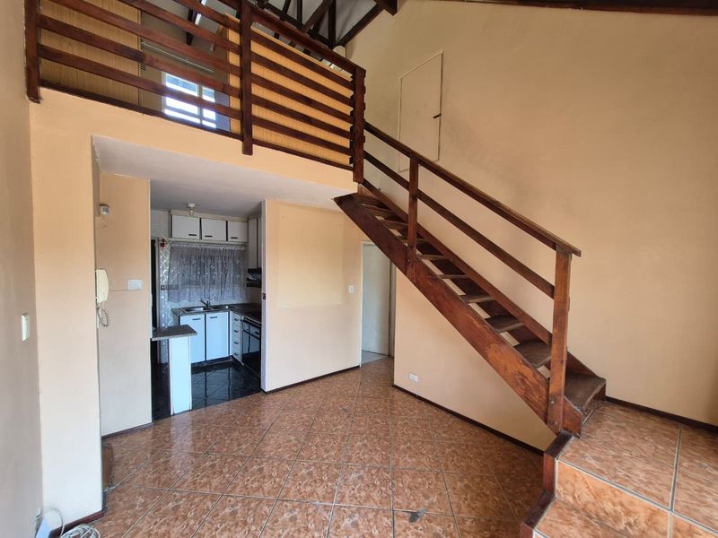 Open plan living area featuring a wooden staircase leading to a mezzanine level, adjacent to a compact kitchen with built-in cabinetry and tiled flooring.