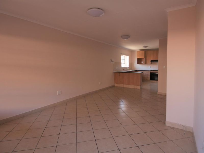 An open-plan living and kitchen area featuring neutral tiled flooring, built-in wooden cabinetry, and integrated kitchen appliances in a clean, vacant space.