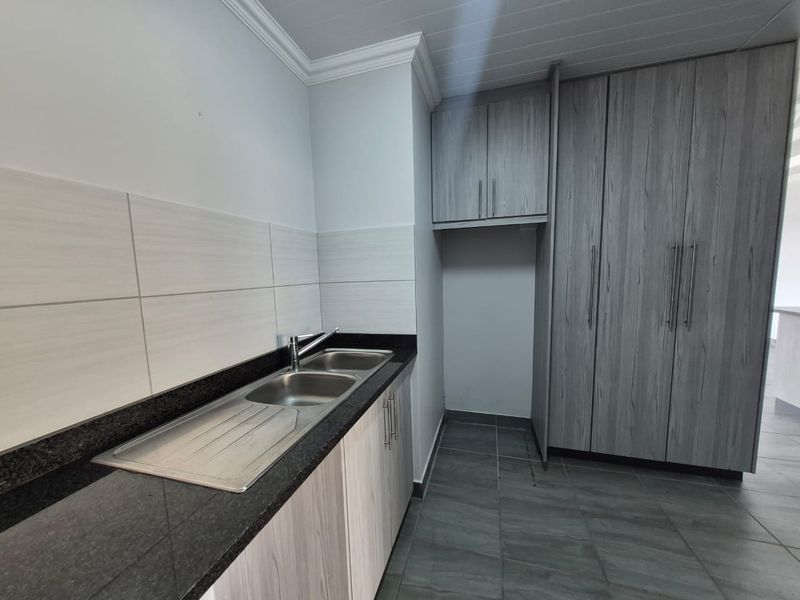 A clean and functional scullery featuring modern grey cabinetry, granite countertops, a double stainless steel sink, and neutral wall tiling.