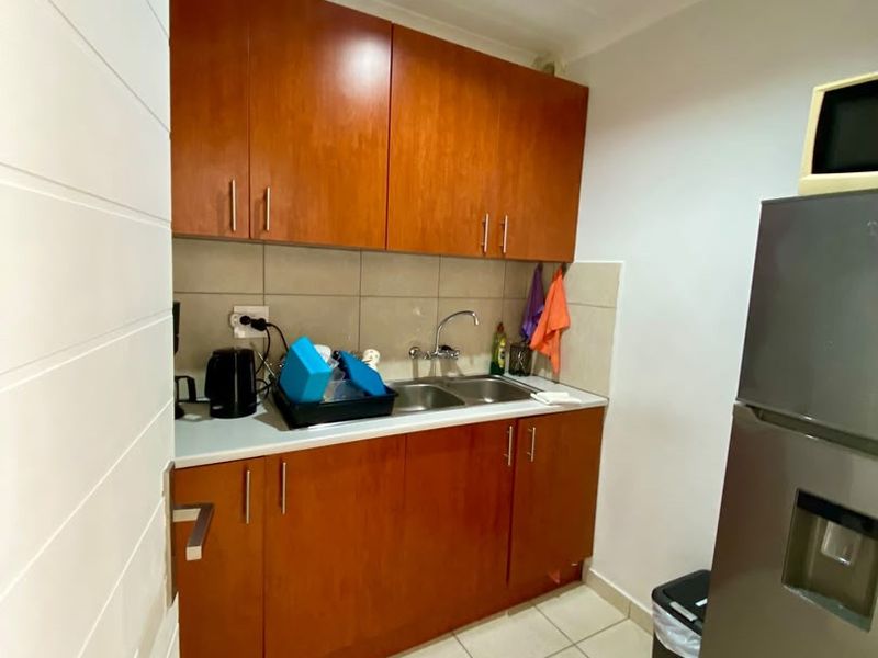 A functional scullery area featuring wooden cabinetry, a stainless steel sink, tiled backsplash, and neutral floor tiles.