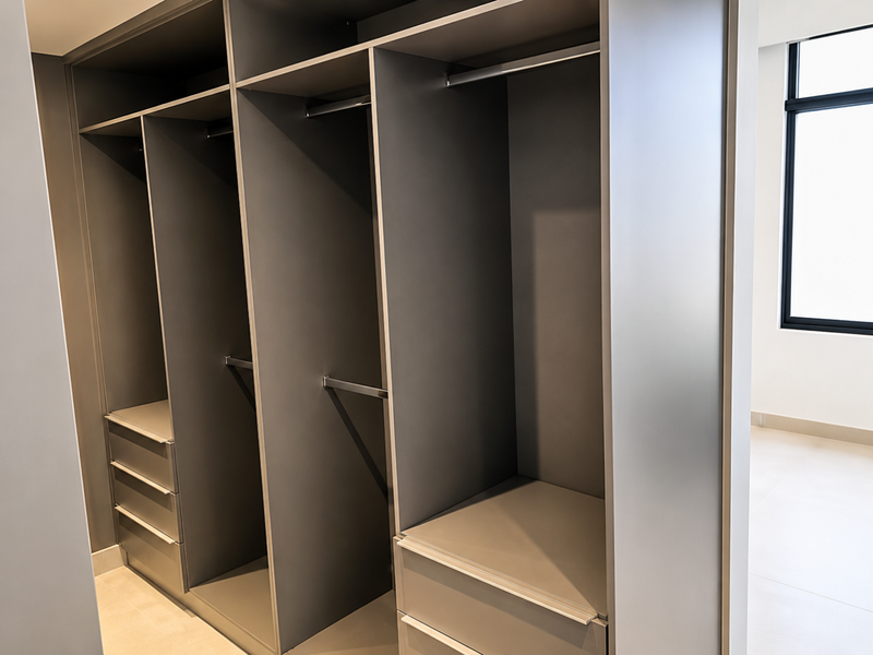 A modern, empty bedroom featuring high-quality built-in cabinetry with integrated hanging rails and drawers, complemented by neutral tiled flooring and a large window.
