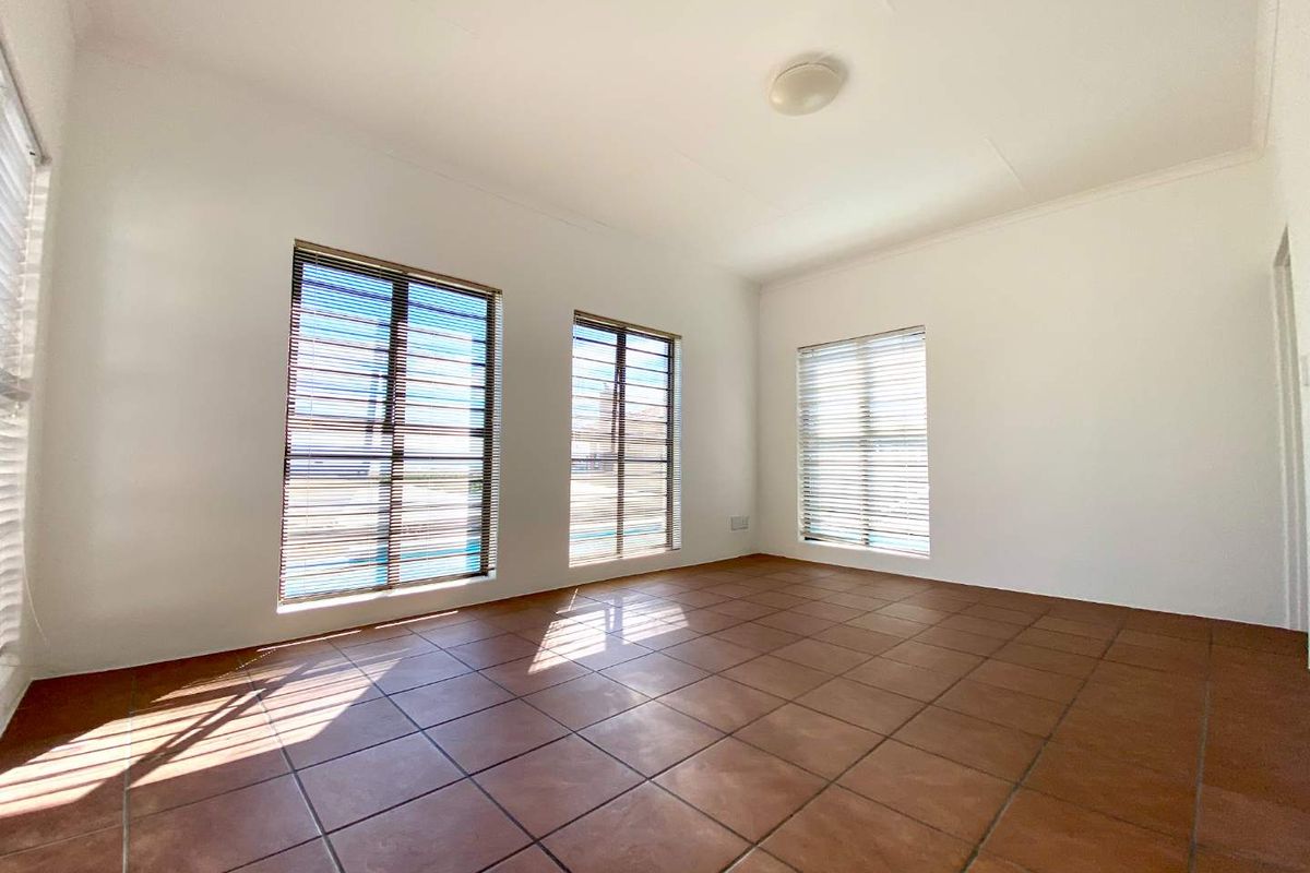 A bright, empty living room featuring terracotta-style floor tiles, large windows with security bars, and fitted venetian blinds.