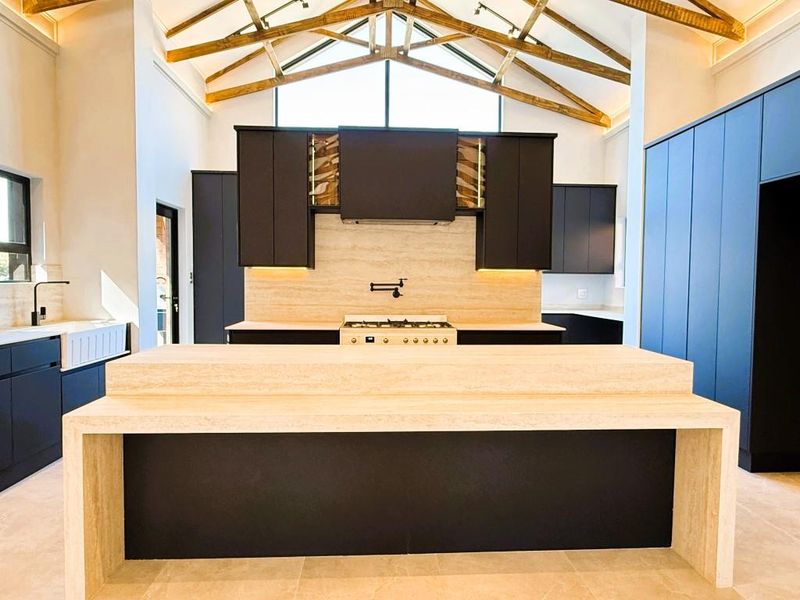 Modern kitchen featuring a large travertine island, farmhouse sink, gas cooktop, and striking exposed timber roof trusses in a high-ceiling open space.