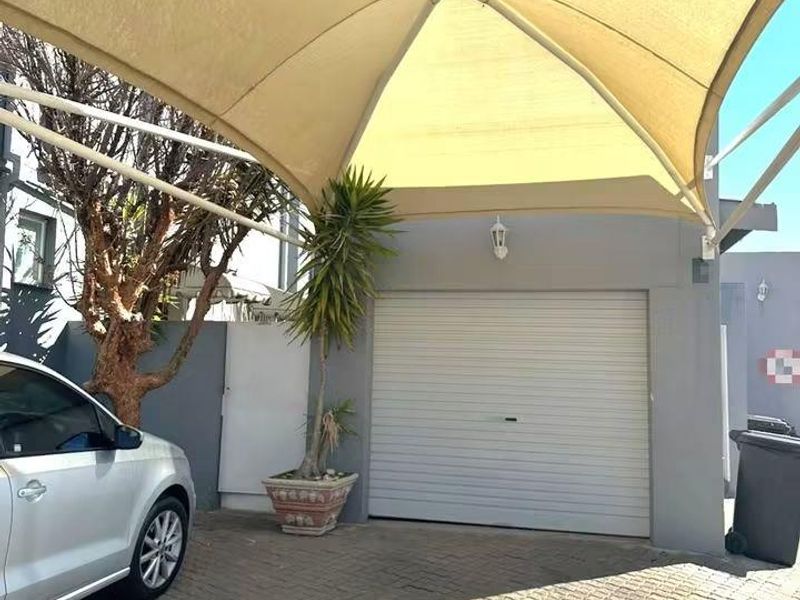 Front exterior view of the townhouse featuring a paved driveway, a secure roller garage door, and a large overhead shade sail structure.