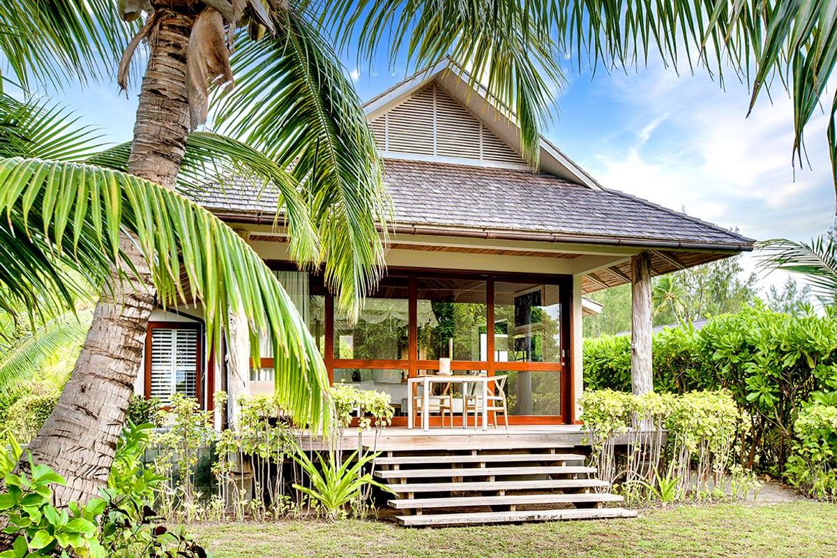 A tropical villa exterior featuring a timber deck, large sliding glass doors, and a pitched shingle roof set within a lush garden.