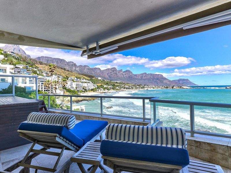 Spacious covered balcony featuring stone flooring, a glass balustrade, and a retractable awning, offering panoramic ocean and mountain views.