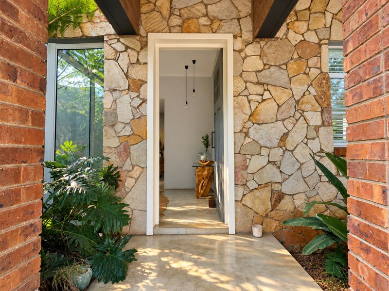 Inviting front entrance featuring natural stone cladding, exposed timber beams, and a polished concrete walkway framed by brick pillars.