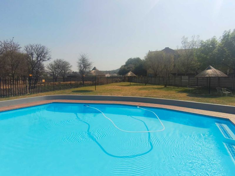 A communal swimming pool area featuring a clear blue pool, paved surround, and landscaped garden space with thatched umbrellas.