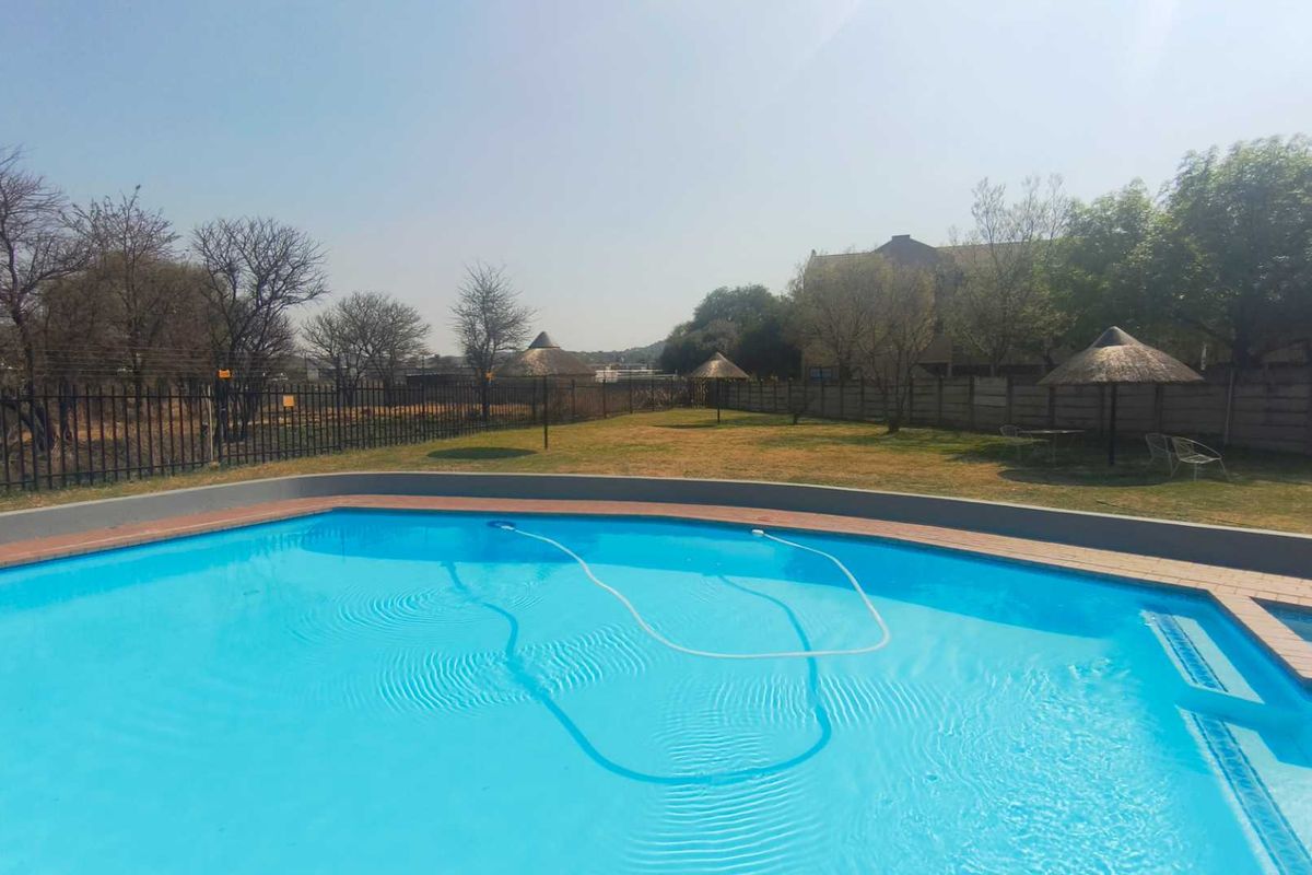 A communal swimming pool area featuring a clear blue pool, paved surround, and landscaped garden space with thatched umbrellas.