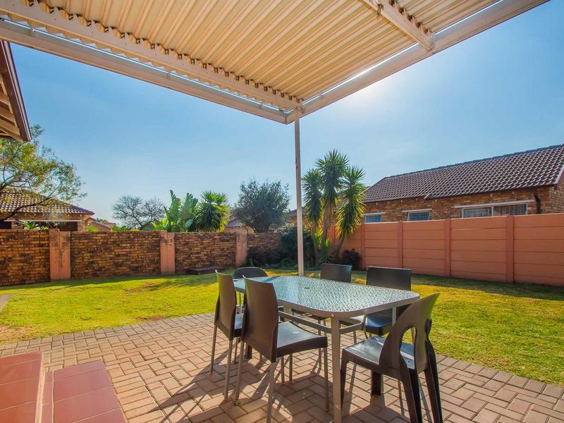 A covered outdoor patio area featuring paved flooring and a corrugated metal roof, overlooking a private garden with brick and precast boundary walls.
