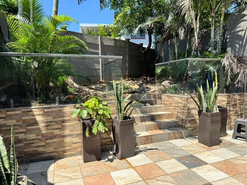 A well-maintained outdoor patio area featuring stone-clad retaining walls, glass balustrades with stainless steel railings, and tiled steps leading to a landscaped garden.